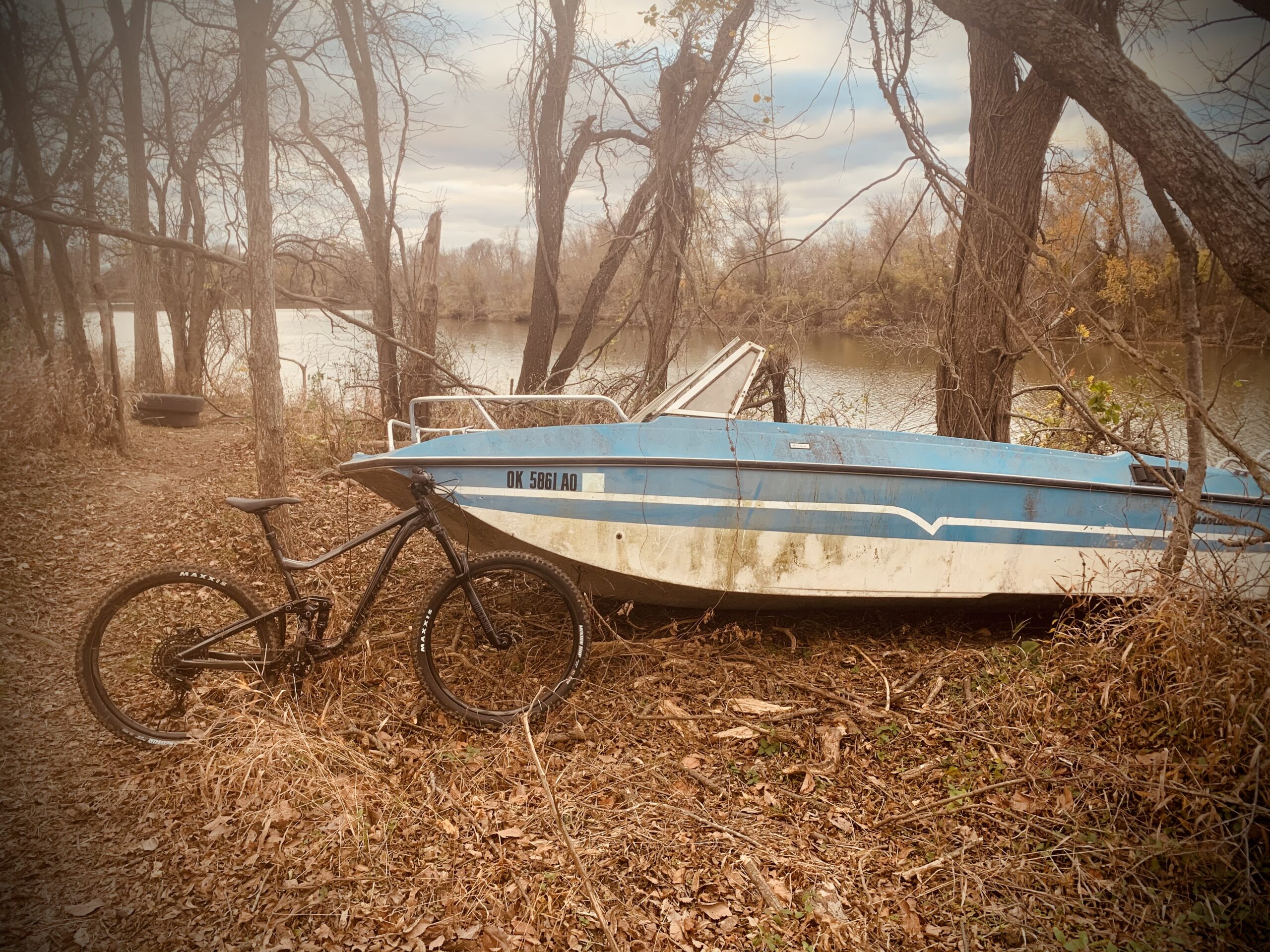 A black mountain bike leaning against a weathered blue boat, partially submerged in a natural setting with bare trees and fallen leaves. The scene reflects a tranquil riverside environment under a cloudy sky. Three Forks Trail mountain bike trail.