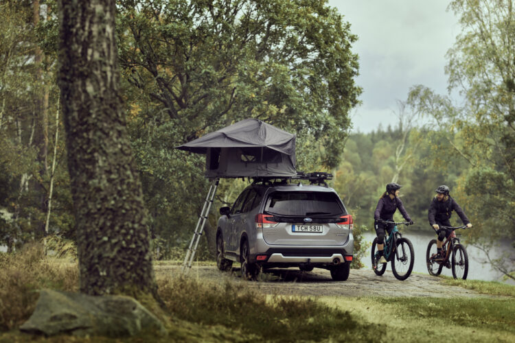 A gray SUV with a rooftop tent parked by a serene lakeside, surrounded by trees. Two mountain bikers in helmets and outdoor gear are riding nearby, engaged in conversation. The scene captures a sense of adventure and outdoor exploration.