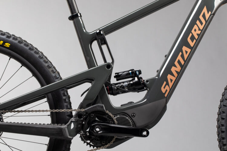 Close-up view of a modern mountain bike frame featuring a gray finish with the brand name "SANTA CRUZ" in orange. The image showcases the bike’s suspension system, chain, and a large, rugged tire, highlighting its durable design suitable for off-road riding. The background is neutral to emphasize the bike's details.