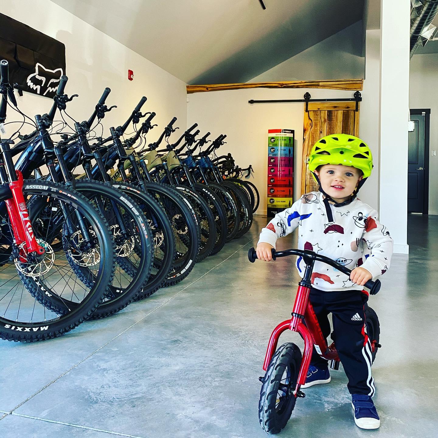 A young child wearing a bright yellow helmet smiles while riding a small red balance bike in a modern bike shop. Behind them, a row of mountain bikes in various colors is neatly lined up, showcasing their tires and handlebars. The interior features a clean, contemporary design with a smooth concrete floor and simple décor.
