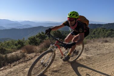 A mountain biker wearing a bright green helmet and a black jersey rides on a sandy trail with a mountainous landscape in the background. The cyclist is in motion, leaning into a turn, with dust kicking up from the rear wheel and a clear blue sky above. Ponti Ridge Trail mountain bike trail.