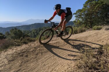 A cyclist in a red shirt and helmet rides a mountain bike along a sandy trail, surrounded by greenery and rolling hills under a clear blue sky. The cyclist leans into the turn, casting a long shadow on the trail. Ponti Ridge Trail mountain bike trail.