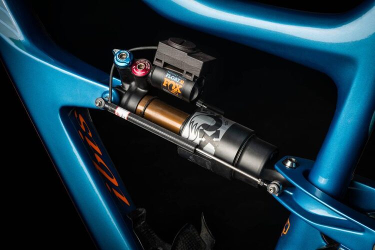 Close-up image of a blue bicycle frame featuring a FOX Float shock absorber. The shot highlights the shock's adjustable features and intricate design, set against a dark background to emphasize the components.