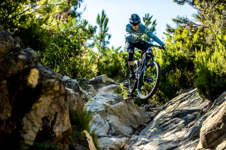 A mountain biker skillfully navigating a rocky trail, airborne over a large rock, surrounded by lush greenery and sunlight filtering through the trees.