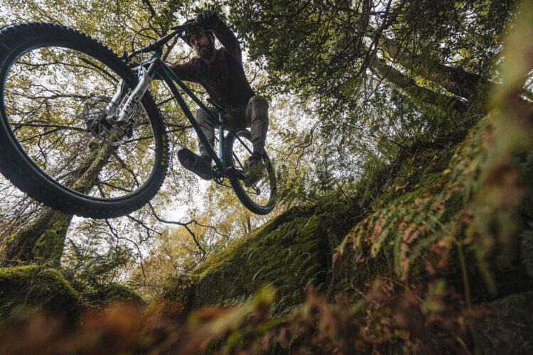Mountain biker performing a jump over rocky terrain, surrounded by trees and foliage, with a dynamic upward perspective.