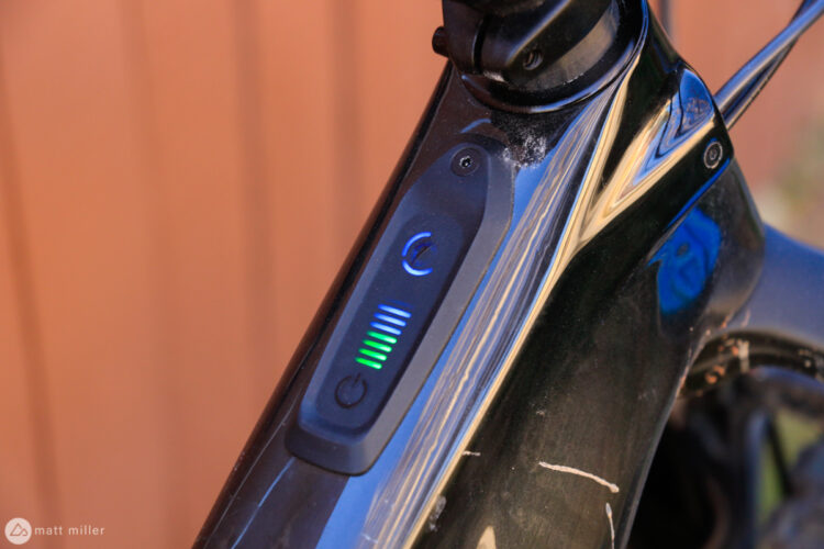 A close-up view of an electric bicycle's control panel, featuring a power button and a battery level indicator with illuminated bars showing different charge levels. The panel is mounted on the frame of the bike, which has a shiny black finish. A soft-focus background highlights the bike's design.