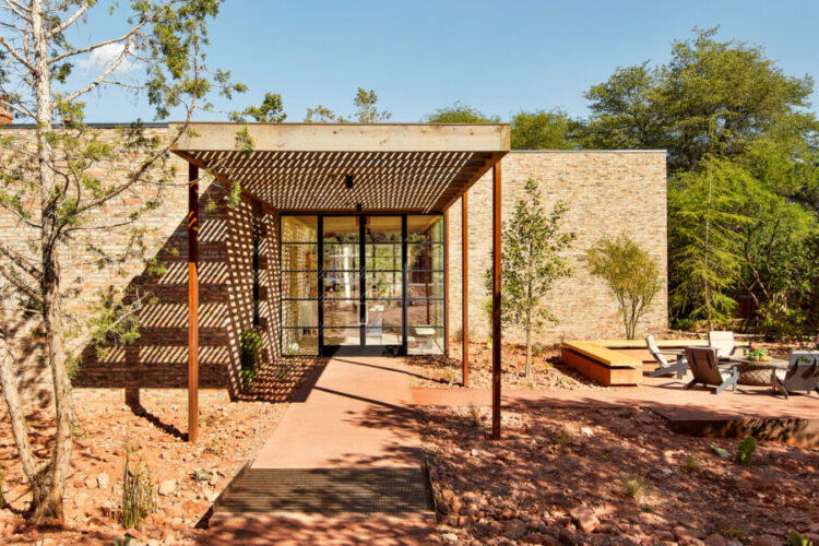 A modern architectural entrance with a sleek glass door framed by horizontal wooden slats. Surrounding the entrance are natural elements, including small trees and aromatic plants set on a reddish-brown landscape. In the background, a patio area features lounge chairs and a wooden platform, all under a clear blue sky.