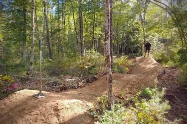 A forested area featuring a dirt bike jump under construction. A shovel stands upright on a pile of dirt, while colorful flags mark the site. In the background, a person stands on a finished jump, surrounded by trees and natural greenery.  Arundel Community Trails mountain bike trail.