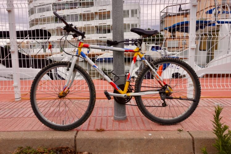 A colorful bicycle with a white frame is parked next to a metal post. The bike features red, blue, and yellow accents and has thick, textured tires suitable for various terrains. In the background, there are boats and a white building with balconies, visible through a white fence. The scene appears to be set near a waterfront area on a cloudy day.