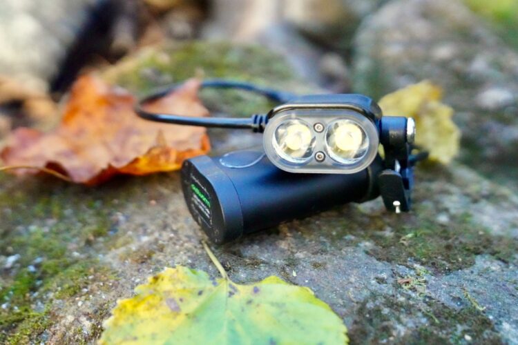 A compact black flashlight with dual LED lights resting on a mossy stone surface, surrounded by autumn leaves in shades of yellow and brown.