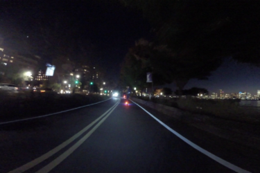 A dimly lit urban road at night, featuring two lanes lined with bright white lane markings. Green traffic lights illuminate the surroundings, while buildings with windows aglow are visible on the left. A cyclist is riding ahead, leaving a trail of light, and the skyline of a city is faintly seen in the distance against the night sky. West Street Greenway mountain bike trail.