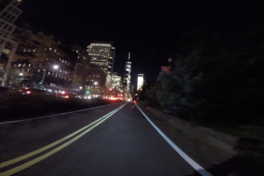 A nighttime view of a city street with illuminated skyscrapers in the background. The scene features a bike lane and double yellow lines, lined with trees on one side and traffic on the other, creating a vibrant urban atmosphere. West Street Greenway mountain bike trail.