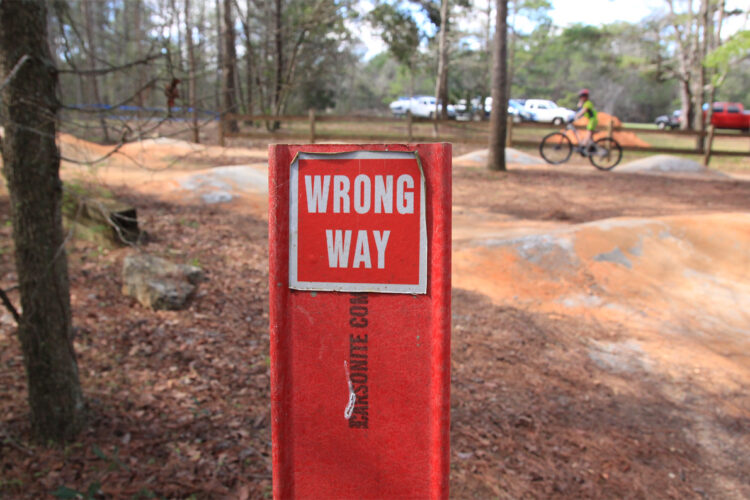 A close-up of a red sign displaying the words "WRONG WAY" positioned among trees in a wooded area. In the background, a cyclist rides along a path near a dirt mound, with parked cars visible further back. The ground is covered in fallen leaves.