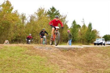 A group of mountain bikers participating in a race on a dirt path surrounded by trees. One young cyclist in a red shirt is jumping over a small rise, while two other riders, dressed in black and blue, are navigating the course behind him. A spectator is watching from the side, and vehicles are parked in the background. The scene is set on a cloudy day. Springhill Park mountain bike trail.