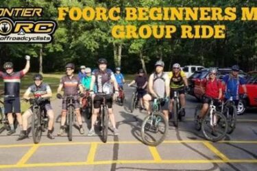A group of mountain biking enthusiasts poses together at the start of a beginners' ride, showcasing a variety of bicycles and riding gear. They stand in a line on a paved area, with several trees in the background, while some participants cheer and display enthusiasm. The image includes the text "FOORC Beginners MTB Group Ride" along with a logo for the Ozark Off Road Cyclists. Springhill Park mountain bike trail.