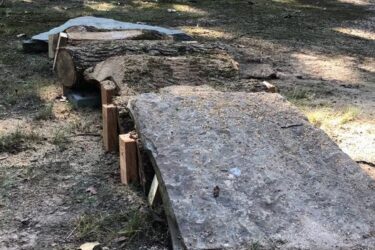 A partially assembled wooden structure and stone slab set in a wooded area, with trees in the background and a car parked on a pathway nearby. The ground is uneven and covered with dirt and grass. Springhill Park mountain bike trail.