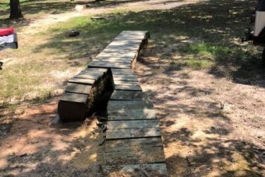 A winding wooden walkway made of planks leads through a sunlit forest area, surrounded by trees and grassy ground. In the background, a glimpse of vehicles can be seen parked along a road. The walkway is partially shaded, emphasizing the natural, rustic charm of the setting. Springhill Park mountain bike trail.