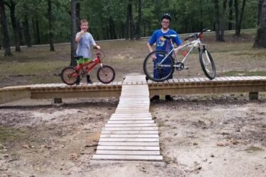 Two boys are standing on a wooden bike bridge in a wooded area. One boy is on a red bike and smiling, while the other, wearing a helmet and a blue shirt, is next to a white bike. The surrounding ground is sandy, and there are trees in the background. Springhill Park mountain bike trail.