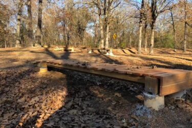 A wooden bridge spans across a small gap in a forested area, surrounded by autumn foliage. The ground is covered with fallen leaves, and the sun filters through the trees, casting shadows on the pathway. In the background, a winding trail is visible, indicating a natural setting for outdoor activities. Springhill Park mountain bike trail.