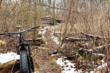 A mountain bike rests against a log on a narrow, winding trail through a winter forest. Bare trees and scattered fallen leaves surround the path, which is partially covered in patches of snow. The scene evokes a calm, natural setting perfect for outdoor exploration. Pinery Provincial Park mountain bike trail.