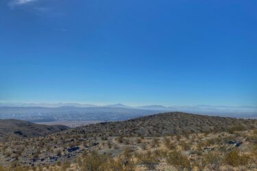 A panoramic view of a rugged desert landscape under a clear blue sky, with the sun shining brightly in the upper left corner. The foreground features rocky terrain and sparse vegetation, while rolling hills and distant mountains create a scenic horizon. Wayne's World mountain bike trail.