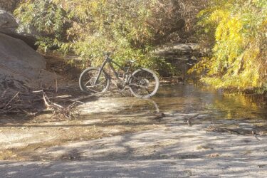 A mountain bike leaning against a rock in a serene outdoor setting, surrounded by trees with autumn foliage. In the foreground, part of another bike's wheel is visible. A small stream flows through the area, creating a peaceful, natural atmosphere. Sycamore Canyon Park mountain bike trail.