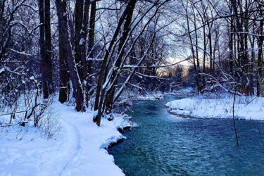 A serene winter landscape featuring a winding river surrounded by bare trees, with a snowy path along the bank. The scene is tranquil, showcasing a light blue sky and reflections on the water's surface. Snow covers the ground and branches, creating a peaceful, frosty atmosphere. Western University trails mountain bike trail.