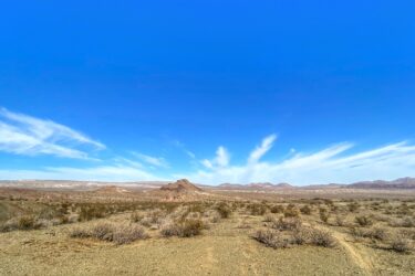 A panoramic view of a desert landscape under a bright blue sky, featuring scattered shrubs and rocky formations in the foreground, with distant mountains and wispy clouds on the horizon. Wayne's World mountain bike trail.