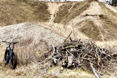 A black fat tire bicycle is parked beside a pile of sticks and branches in an open field. In the background, there are grassy hills with dirt trails winding up their slopes. The sky is overcast, contributing to a gray, muted atmosphere. Hardy Rd. Trail mountain bike trail.