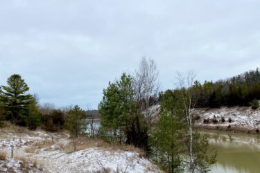 A serene landscape featuring a partially snow-covered pathway leading to a calm body of water. The scene includes a mix of evergreen and deciduous trees, with a cloudy sky overhead. The foreground shows patches of grass and a small tree, while the water reflects the surrounding natural scenery. Lambton County Heritage Forest mountain bike trail.