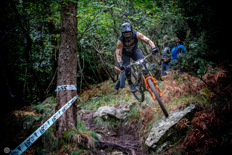 A mountain biker in full protective gear is airborne while navigating a rocky trail in a wooded area. The rider, with a black helmet and gloves, is moving swiftly over a terrain marked by trees and ferns, with a race number visible on their bike. An audience member can be seen in the background, adding to the dynamic atmosphere of the scene.