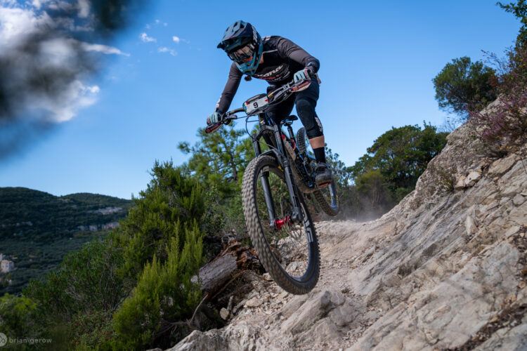 A mountain biker jumping off a rocky trail, surrounded by trees and hills under a blue sky. Dust is kicking up from the trail as the rider maneuvers through the terrain, showcasing action and adventure in mountain biking.