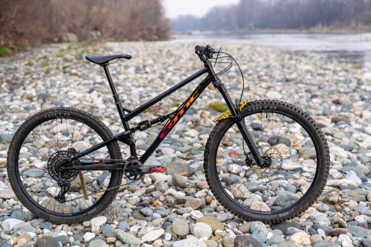 A black mountain bike with orange and pink accents resting on a rocky riverbed. The bike features thick tires designed for off-road use, a prominent seat post, and a sleek frame, surrounded by a variety of smooth pebbles and stones with a blurred river in the background.