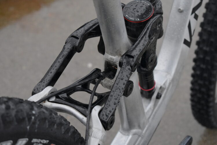 Close-up view of a bicycle's rear suspension system, featuring a combination of black and silver components with water droplets, indicating a wet environment. The image highlights the intricate mechanics of the suspension linkage and shock absorber.
