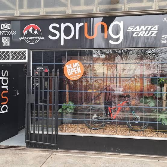 Storefront of a bike shop named "Sprung" featuring logos of various brands such as Rocky Mountain and Santa Cruz. A bicycle is displayed prominently in the window along with indoor plants. A sign stating "WE ARE OPEN" is visible. The overall design is modern with a black and orange color scheme.