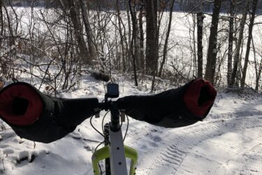 A bicycle equipped for winter riding in a snowy landscape, featuring large handlebar mitts for warmth. The view is from the rider's perspective, showing a snow-covered path and a frozen lake in the background, surrounded by trees. The sky is clear with a hint of sunlight. Standing Rocks mountain bike trail.