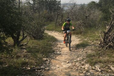 A cyclist riding a mountain bike along a rocky trail in a natural landscape, surrounded by sparse trees and shrubs under a cloudy sky. Dana Peak mountain bike trail.