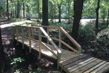 Wooden footbridge crossing a small creek in a forested area, surrounded by trees and underbrush. Sunlight filters through the leaves, casting dappled shadows on the bridge. Springhill Park mountain bike trail.