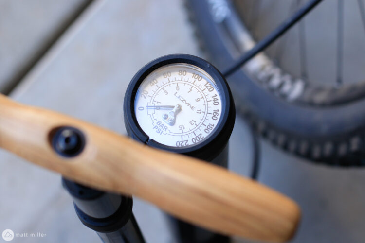 Close-up of a bike pump gauge displaying pressure measurements in PSI and BAR, with a wooden handle. A bicycle wheel is partially visible in the background.