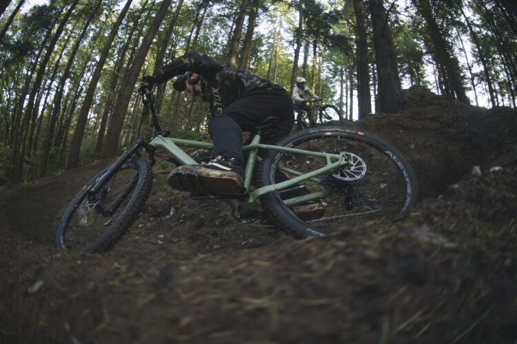 A mountain biker navigating a dirt trail in a forest, leaning into a curve on a green bicycle, with another biker visible in the background. The scene is surrounded by tall trees and dappled sunlight filtering through the leaves.
