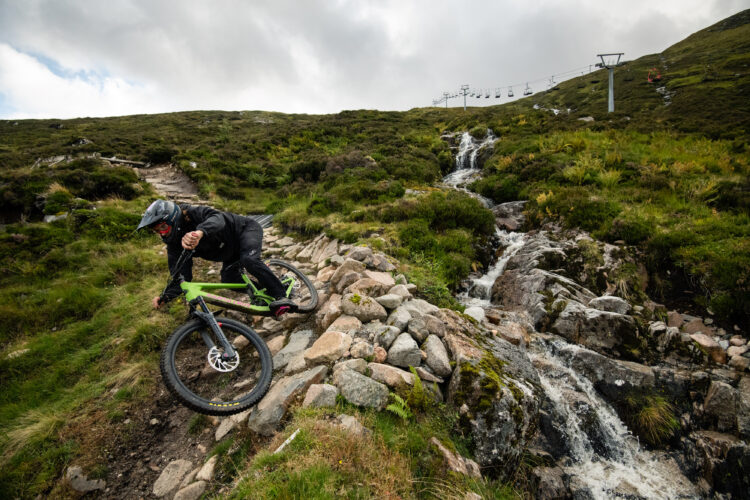 A mountain biker navigating a rocky terrain next to a stream, wearing a helmet and black gear. In the background, a ski lift is visible against a cloudy sky. The landscape features lush greenery and rugged rocks.
