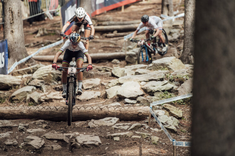 Three mountain bikers navigate a rocky trail, featuring logs and uneven terrain in a wooded area. The foreground shows a rider in a white jersey with colorful stripes, focused and balancing on their bike. Two additional riders are visible in the background, also maneuvering through the challenging course. Bright banners and fencing line the trail, typical of a competitive mountain biking event.