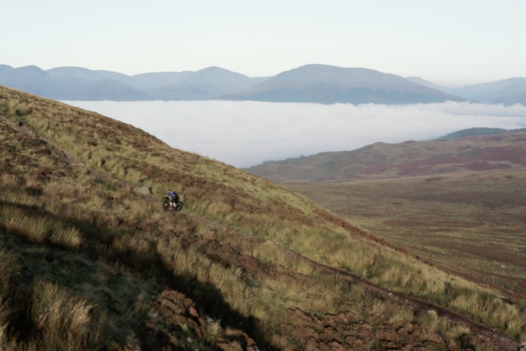 A cyclist navigates a rugged hillside trail, surrounded by lush green grass and rocky terrain, with misty mountains and clouds visible in the background under a clear sky.