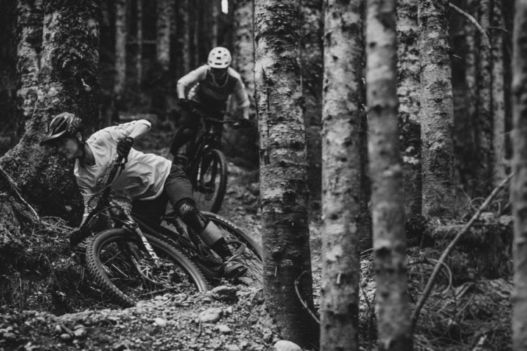 Two mountain bikers navigate a winding trail through a dense forest, with tall trees surrounding them. The image is in black and white, capturing the dynamic movement of the riders as they lean into the turns, showcasing the rugged terrain and their sporty attire.