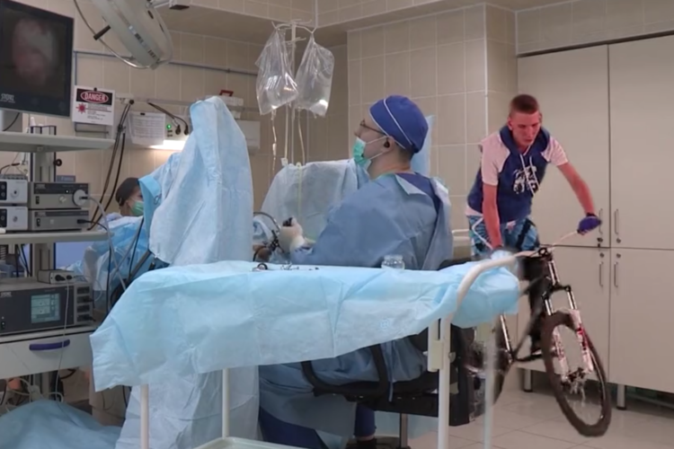 A hospital operating room with medical staff in blue scrubs and masks attending to a patient. A young man on a bicycle is riding through the room. Medical equipment, including monitors and IV bags, is visible in the background.