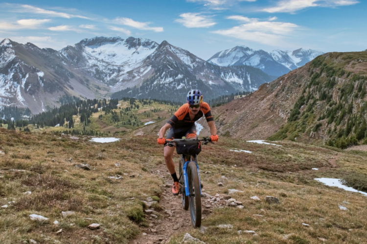 A mountain biker navigates a rocky trail surrounded by snow-capped mountains and lush greenery under a blue sky with scattered clouds. The cyclist wears a helmet and an orange jersey, focused on riding through the rugged terrain.