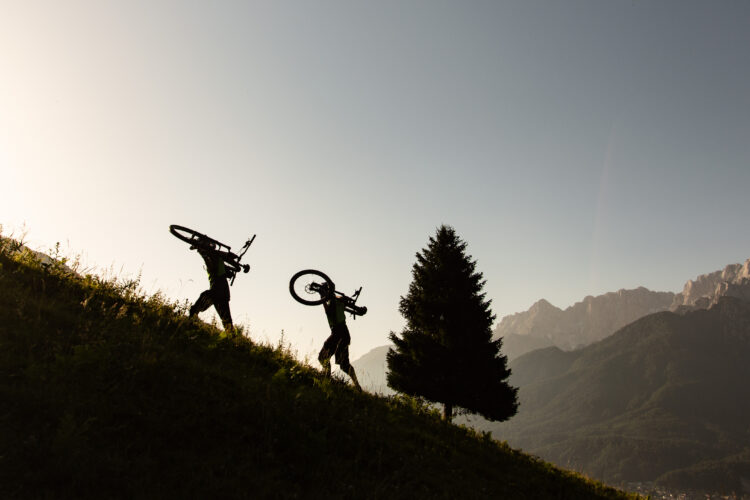 Two mountain bikers silhouetted against a sunset, walking uphill while carrying their bicycles. A lone tree stands nearby with a mountainous landscape in the background.