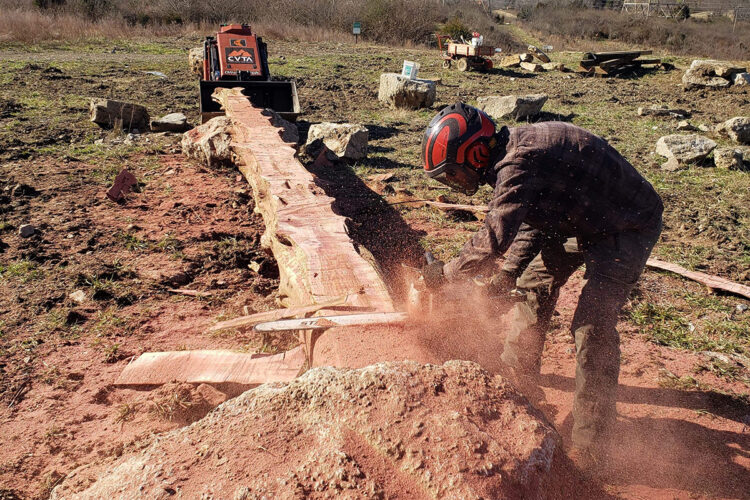 A person wearing a helmet and protective gear uses a chainsaw to cut a large log on a construction site. Dust and wood shavings fly into the air as they work, with a trailer and equipment visible in the background on a grassy, uneven terrain.