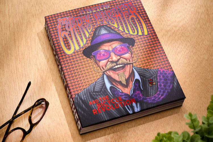 An artistic book cover featuring a smiling man with a mustache, wearing sunglasses and a purple hat. The background has a vibrant, patterned design, and the title reads "Eary Fisher and the Bicycle Revolution." The book is placed on a wooden surface alongside a pair of eyeglasses and a small green plant.