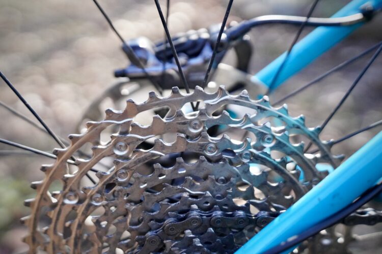 Close-up of a bicycle's rear cassette gears, featuring multiple metal sprockets arranged in a circular pattern, with a blurred background and part of the bike's frame visible in light blue.